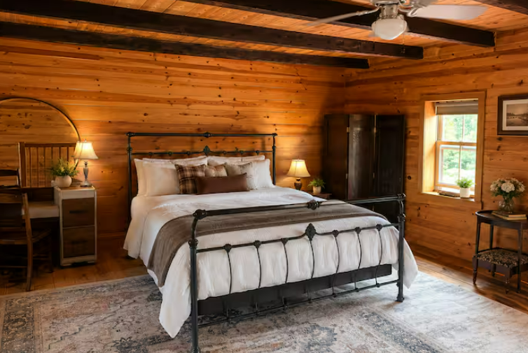 Bedroom with a black iron bed, plaid pillows, log walls, ceiling fan, and warm lamplight