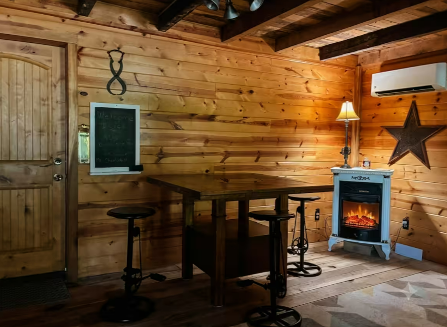 Dining nook with wooden table and stools, chalkboard welcome sign, barn star, and a glowing fireplace stove